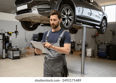 Mechanic using digital tablet in car repair shop with raised vehicle in background. Man in work uniform checking diagnostics. Auto service, modern technology in automobile maintenance. - Powered by Shutterstock