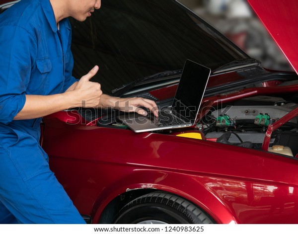 mechanic using laptop computer to check a car\
engine and showing thumbs up at the repair garage. Auto repair, car\
service and maintenance\
concept.