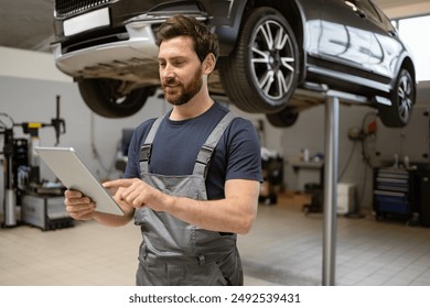 Mechanic using tablet in car repair shop under lifted vehicle. Focused automotive technician inspecting diagnostics, modern technology in auto maintenance, professional service environment. - Powered by Shutterstock