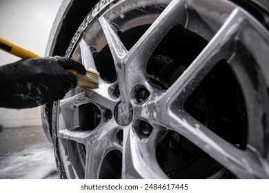 Mechanic wearing black gloves cleaning a car wheel with a brush in a detailing studio. Close-up view of hands scrubbing the wheel rim to remove dirt and grime, ensuring a spotless finish - Powered by Shutterstock