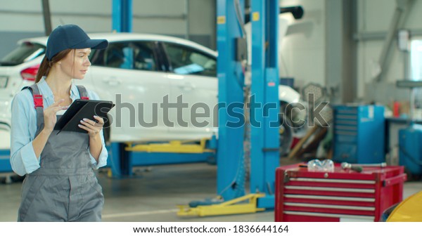 Mechanic woman with\
tablet walking along car workshop. Car service, repair, maintenance\
and people concept.