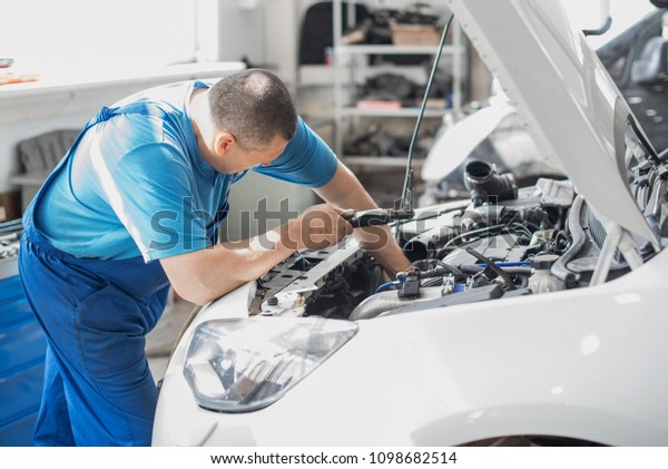 A mechanic working in\
a car workshop in a blue working form is repairing a gray car. Auto\
service.