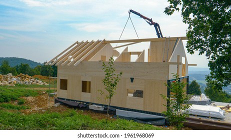 Mechanical beam lifts a wooden roofing beam to the top floor of a prefabricated house under construction in the lush green countryside. Rural landscape surrounds a CLT house being built atop a hill.