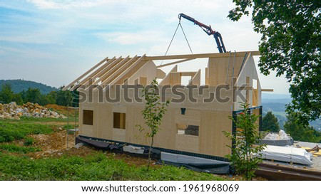 Mechanical beam lifts a wooden roofing beam to the top floor of a prefabricated house under construction in the lush green countryside. Rural landscape surrounds a CLT house being built atop a hill.