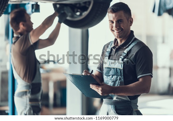 Mechanics Checking Wheel Bearings in Car\
Workshop. Two Caucasian Adult Technician Master in Uniform\
Repairing Car. Repairman Looking at Camera Filling Clipboard of\
Service Order. Auto Service\
Concept