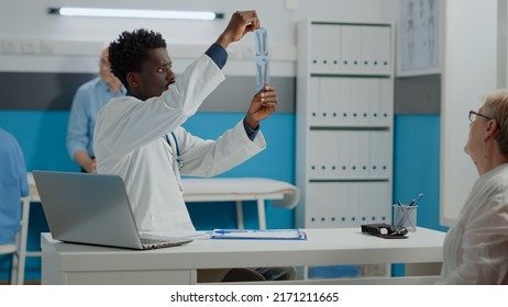 Medic Of African American Ethnicity Analyzing X Ray Of Patient Healthcare Sitting At Desk. Black Doctor Holding Radiography Scan For Professional Diagnosis And Results In Medical Cabinet