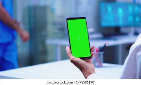 Medic In Hospital Cabinet Holding Phone With Green Screen Wearing White Coat While Nurse Opens Glass Door. Healthcare Specialist In Hospital Cabinet Using Smartphone With Mockup.