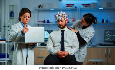 Medic in neuroscience working in neurological research laboratory developing brain experiment holding laptop explaning to man brainwave scanning headset side effects of nervous system treamtment.