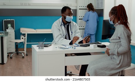 Medic And Pregnant Woman With Face Mask Talking About Pills Treatment In Bottle At Checkup Appointment. Patient With Pregnancy Belly Receiving Prescription Medicine From Doctor In Cabinet.