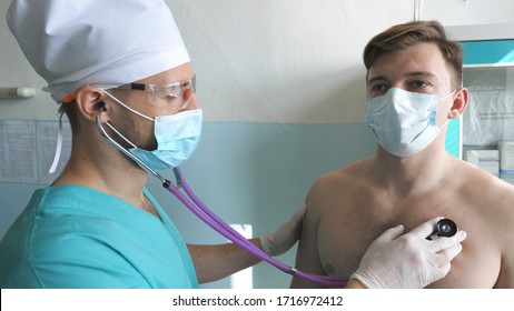 Medic With Stethoscope Listens Lungs Of Patient Looks For COVID-19 Symptoms. Doctor With Examining Man With Stethoscope To Coronavirus Pneumonia Signs. Physician Checking Chest Of Guy At The Hospital.