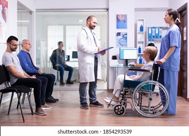 Medic Talking With Disabled Woman In Hospital Waiting Area While Inviting Patient In Examination Room. Young Man Waiting For Doctor Consultation. Health Care System And Medicine Staff