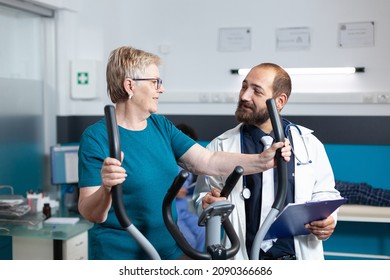 Medic talking to senior patient doing physical activity to recover from injury with electrical bike. Elder woman using stationary bicycle for wellness and recovery at physiotherapy clinic. - Powered by Shutterstock