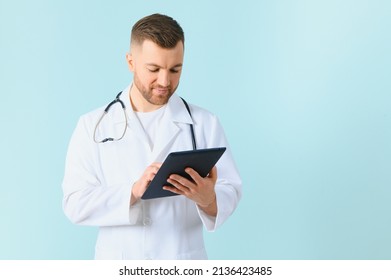 Medic In White Robe, Looking Straight At Camera With Positive Smile, Holding Tablet Computer Next To Chest While Working In Hospital, Isolated On Blue Background