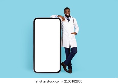 Medical Advertisement. Handsome Doctor In Uniform Standing Near Big Blank Smartphone, Happy Smiling African American Physician Recommending Mobile App Or Website Over Blue Background, Mockup
