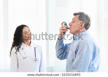 Medical Advice. Elderly man using asthma inhaler, mexican doctor watching, free space