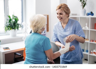 Medical aid. Joyful positive nurse smiling to her patient while wrapping bandage around her hand - Powered by Shutterstock