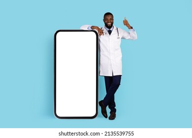Medical App. African American Doctor In Uniform Posing Near Big Blank Smartphone And Gesturing Thumb Up At Camera, Happy Black Male Therapist Standing Over Blue Studio Background, Mockup