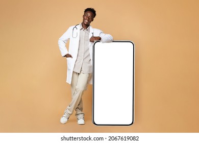 Medical App. African American Male Doctor In Uniform Leaning At Big Blank Smartphone With White Screen, Demonstrating Free Copy Space For Advertisement, Standing On Beige Background, Mockup