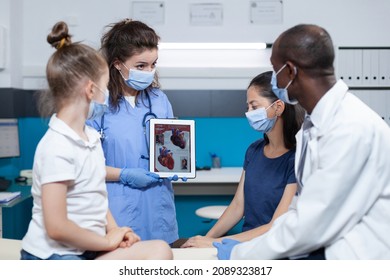 Medical Asisstant Holding Tablet With Heart Expertise On Screen During Clinical Appointment In Hospital Office. African American Pediatrician Doctor With Protective Face Mash Against Coronavirus