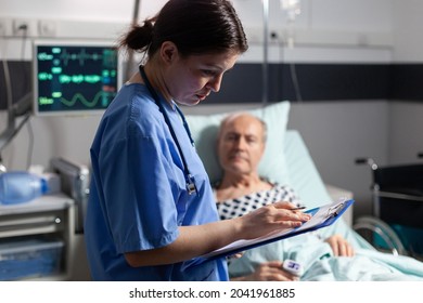 Medical Assistant Checking Treatment Of Senior Man Reading Notes On Clipboard. Sick Elderly Laying In Bed Discussing With Nurse About Diagnosis Expertise With Iv Bag Attached.