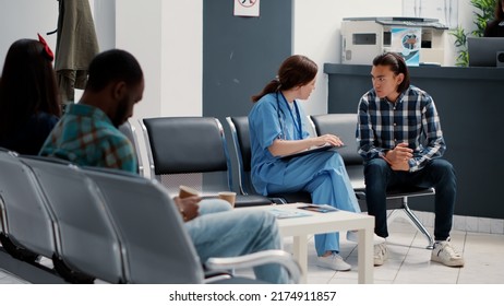 Medical Assistant Filling In Checkup Report And Talking To Asian Patient In Hospital Reception Lobby. Nurse Discussing About Disease Treatment And Medicine Support With Person In Waiting Area.
