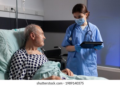 Medical Assistant With Protection Mask Checking Treatment Of Senior Man Reading Notes On Tablet Pc. Sick Elderly Laying In Bed Discussing With Nurse About Diagnosis Expertise With Iv Bag Attached.