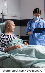 Medical Assistant With Protection Mask Checking Treatment Of Senior Man Reading Notes On Tablet Pc. Sick Elderly Laying In Bed Discussing With Nurse About Diagnosis Expertise With Iv Bag Attached.