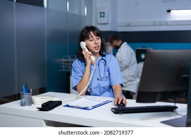 Medical Assistant Talking On Landline Phone For Healthcare Checkup Appointment, Working Late. Woman Nurse Using Telephone For Remote Communication With Patient While Sitting At Desk.