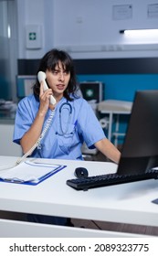 Medical Assistant Talking On Telephone For Checkup Appointment, Working Late. Woman Nurse Using Landline Phone For Online Remote Communication With Patient At Healthcare Cabinet.