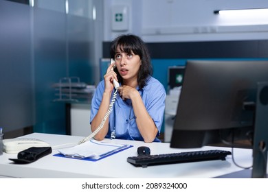 Medical Assistant Talking On Telephone To Patient For Appointment, Working Late. Woman Nurse Using Landline Phone For Remote Communication And Healthcare System While Sitting At Desk.