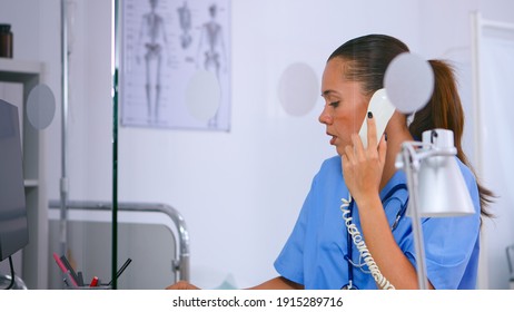 Medical Assistant Talking At Phone And Typing On Computer Offering Consultation In Hospital Clinic. Woman Receptionist In Medicine Uniform, Doctor Nurse Assistant Helping With Telehealth Communication