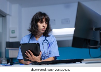 Medical Assistant Using Tablet With Touch Screen For Healthcare System. Nurse Holding Digital Gadget And Looking At Computer, Working Late At Night. Specialist With Modern Device