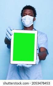 Medical Assistant With Vertical Green Screen On Tablet Looking At Camera. Nurse With Face Mask Holding Device With Chroma Key, Showing Isolated Mockup Template. Healthcare Specialist