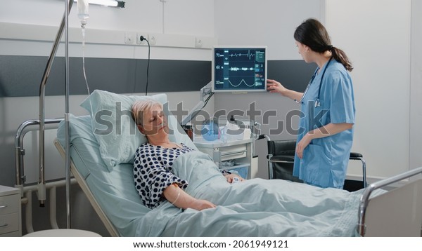Medical\
assistant visiting retired patient sleeping in hospital ward bed.\
Woman working as nurse checking on heart rate monitor for pulse\
measurement. Specialist taking care of\
person