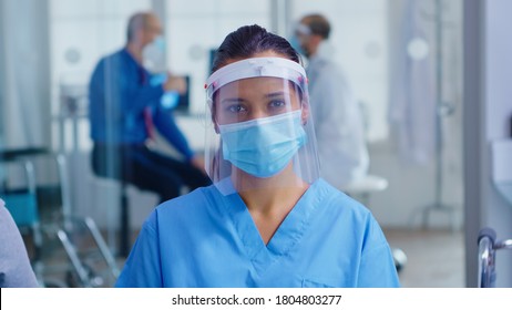 Medical assistant with visor and face mask against coronavirus looking at camera in hospital waiting area. Doctor consulting senior man in examination room.