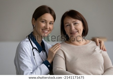 Medical care. Headshot portrait of two females young doctor and elderly patient pensioner on appointment at clinic. Happy confident attending physician hug shoulders of mature lady client help support