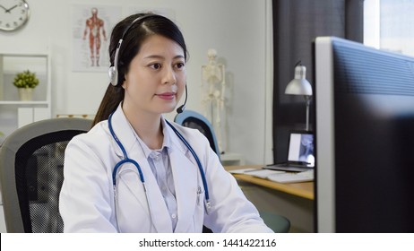 Medical Center Concept. Asian Woman With Headset Working In Clinic Office On Computer At Desk. Smiling Elegant Female Nurse Pick Up Answer Phone Calls By Patient. Girl In Lab Coat And Stethoscope