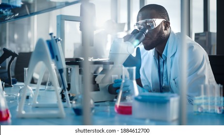 Medical Development Laboratory: Black Male Scientist Looking Under Microscope, Inspecting Petri Dish. Professionals Working In Advanced Scientific Lab Doing Medicine, Vaccine, Biotechnology Research