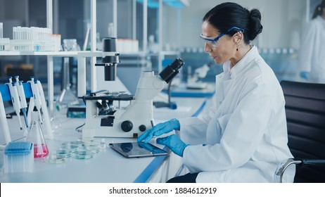 Medical Development Laboratory: Portrait Of Beautiful Caucasian Female Scientist Looking Under Microscope, Enters Data Into Digital Tablet. Medicine, Biotechnology Research In Advanced Pharma Lab