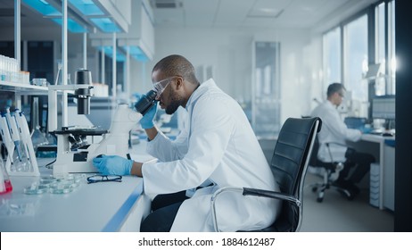 Medical Development Laboratory: Portrait Of Black Male Scientist Looking Under Microscope, Analyzing Petri Dish Sample. Professionals Doing Research In Advanced Scientific Lab. Side View Shot