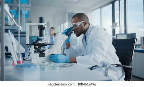 Medical Development Laboratory: Portrait of Black Male Scientist Looking Under Microscope, Analyzing Petri Dish Sample. Professionals Doing Research in Advanced Scientific Lab. Side View Shot