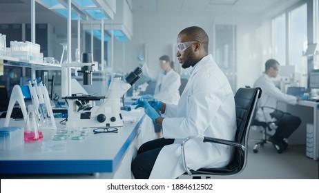 Medical Development Laboratory: Portrait Of Black Male Scientist Looking Under Microscope, Analyzing Petri Dish Sample. Professionals Doing Research In Advanced Scientific Lab. Side View Shot