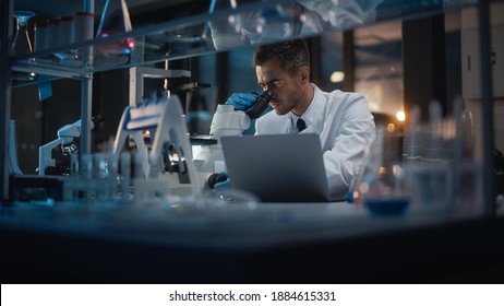 Medical Development Laboratory: Portrait Of Male Scientist Works On Microscope, Using Laptop, Enters Data For Analysis. Pharmaceutical Lab For Medicine, Biotechnology, Drugs, Research. Evening Work