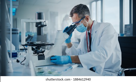 Medical Development Laboratory: Scientist Wearing Face Mask Looking Under Microscope. He Mix Petri Dish Sample. Specialists Working on Medicine, Biotechnology Research in Advanced Pharma Lab
