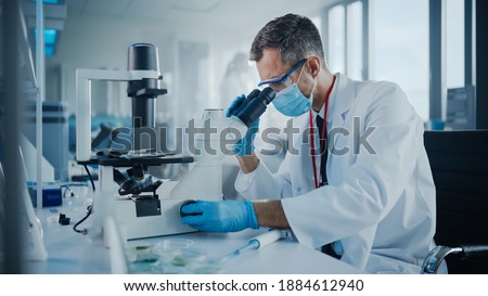 Medical Development Laboratory: Scientist Wearing Face Mask Looking Under Microscope. He Mix Petri Dish Sample. Specialists Working on Medicine, Biotechnology Research in Advanced Pharma Lab