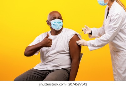 Medical Doctor Giving Coronavirus Vaccine Shot To Senior Black Man Over Orange Background. African American Male In Face Mask Getting Covid-19 Vaccine, Showing Thumb Up, Recommending Immunization