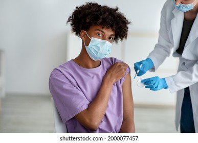 Medical Doctor Or Nurse Giving Coronavirus Vaccine Shot To Black Teenage Guy At Clinic. African American Adolescent In Face Mask Getting Covid Vaccination, Participating In Immunization Campaign