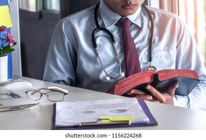Medical Doctor Reading Bible With On Desk.