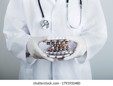 Medical Doctor Wearing Clear Latex Gloves Holding Stack Of Different Pills, Antibiotics And Virus Treatment Tablets On Grey Hospital Wall.