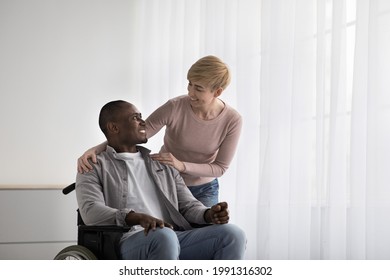 Medical Equipment In Hospital For Assistance Handicapped People And Help. Smiling Woman Nurse Care About Smiling Adult African American Disabled Guy In Wheelchair, In Living Room Or Clinic Interior